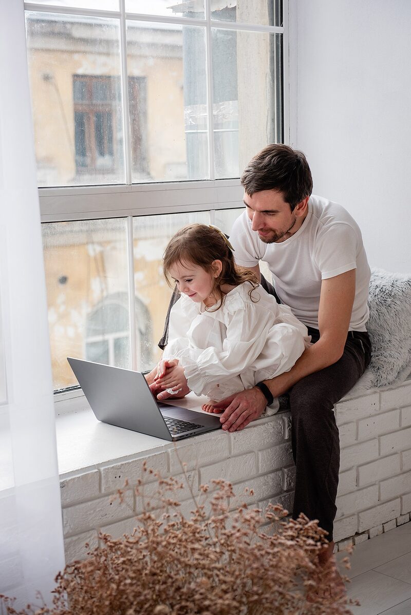 A father and daughter sitting by a sunlit window, learning together on a laptop.